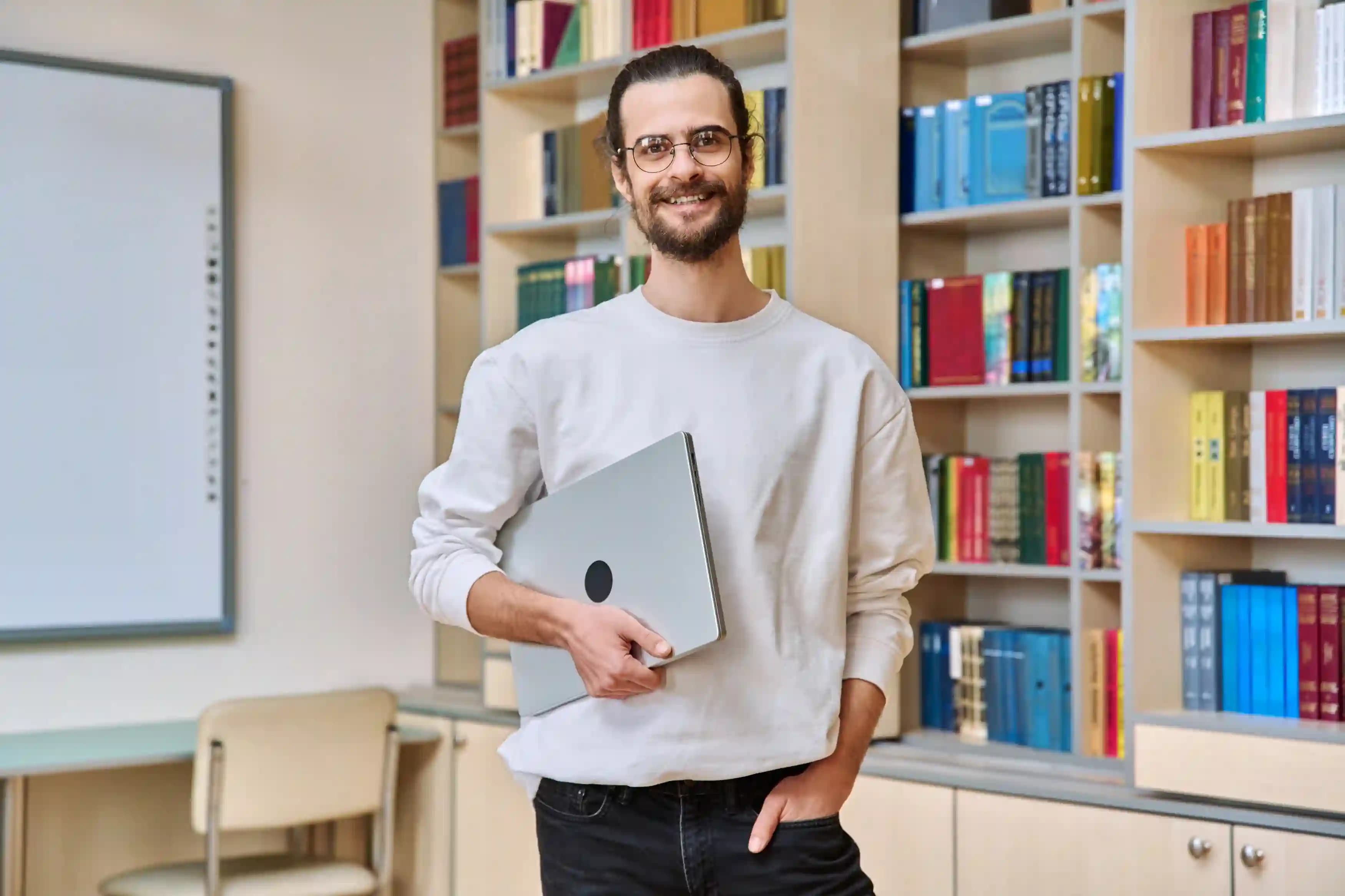 Lehrkraft steht mit zugeklapptem Laptop unter dem Arm vor einem Bücherregal