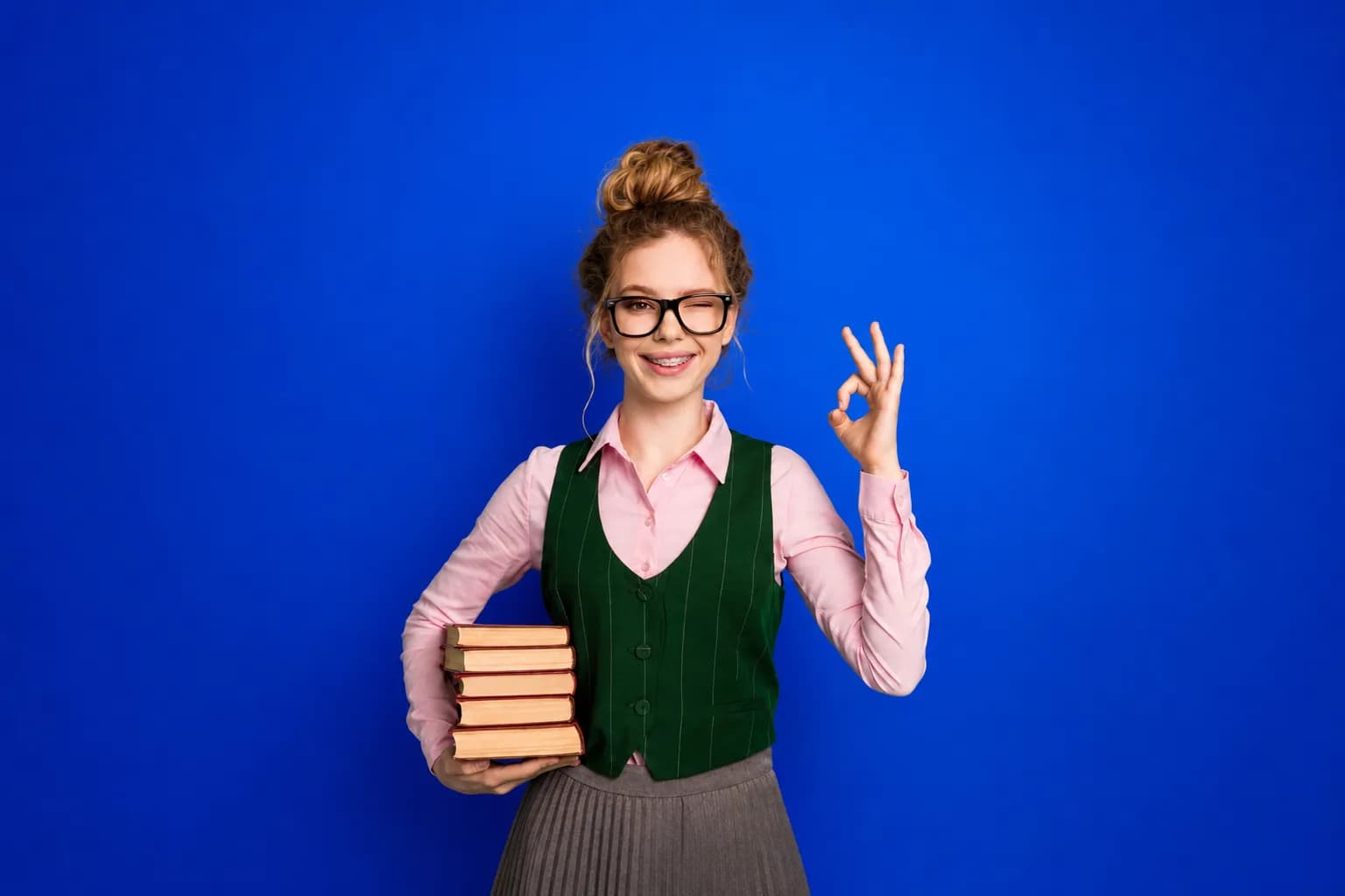 Schülerin mit Büchern unter dem Arm zwinkert und zeigt das Okay-Zeichen